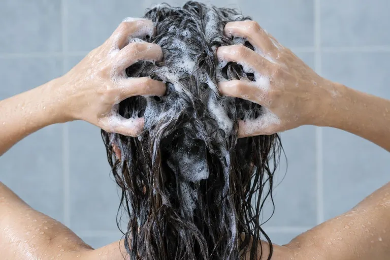 Twee handen masseren schuim in nat donker haar, van achteren gezien, met druppels op de schouders