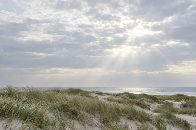 Zon die door een wolkendek breekt boven duinen met helmgras en de zee