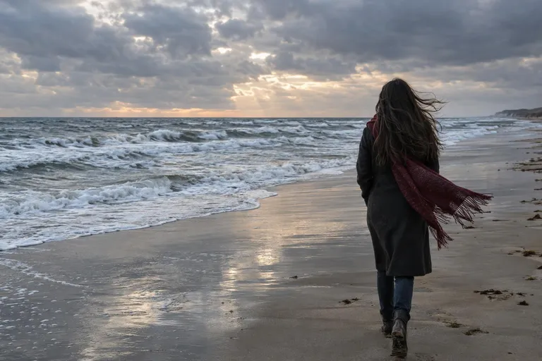 Een vrouw met lang donker haar loopt van ons af over een winderig strand in de herfst, haar en sjaal waaien op