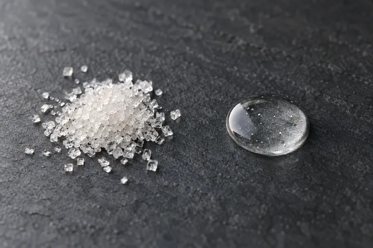 Een hoopje grove suikerkorrels naast een heldere druppel gel op donkere leisteen